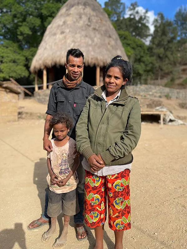 Family of three standing in front of a traditional hut with trees in the background